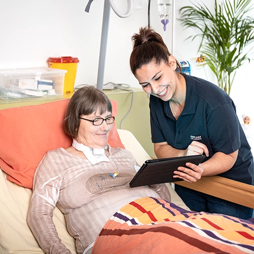 Weibliche Pflegekraft am Bett einer Klientin, die mit hochgefahrener Lehne im Bett sitzt. Die Pflegekraft, etwas vornübergebeugt, zeigt ihr etwas auf einem Tablet.